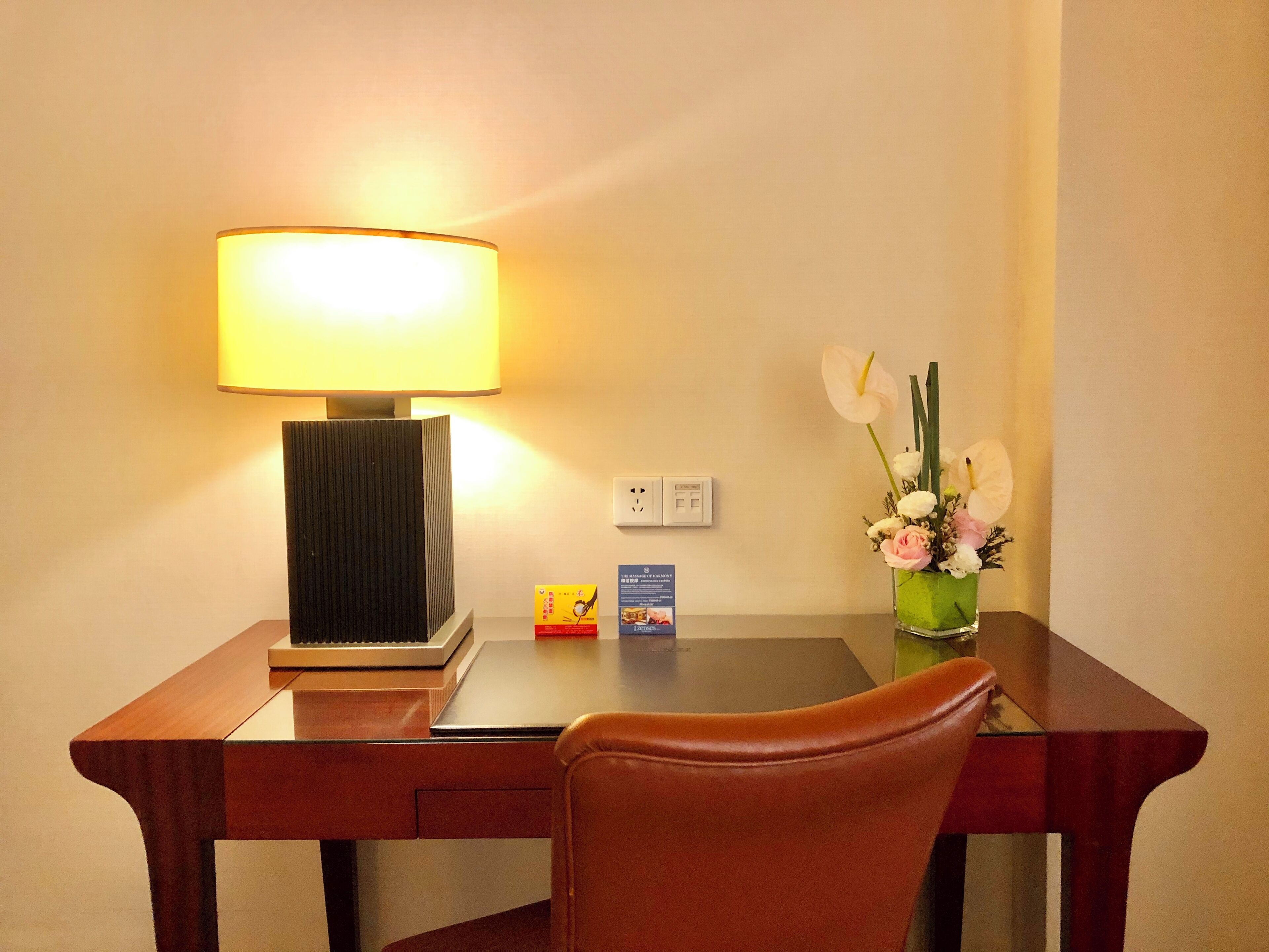 A hotel room desk with a lamp, a vase with flowers, and a small box on the table.