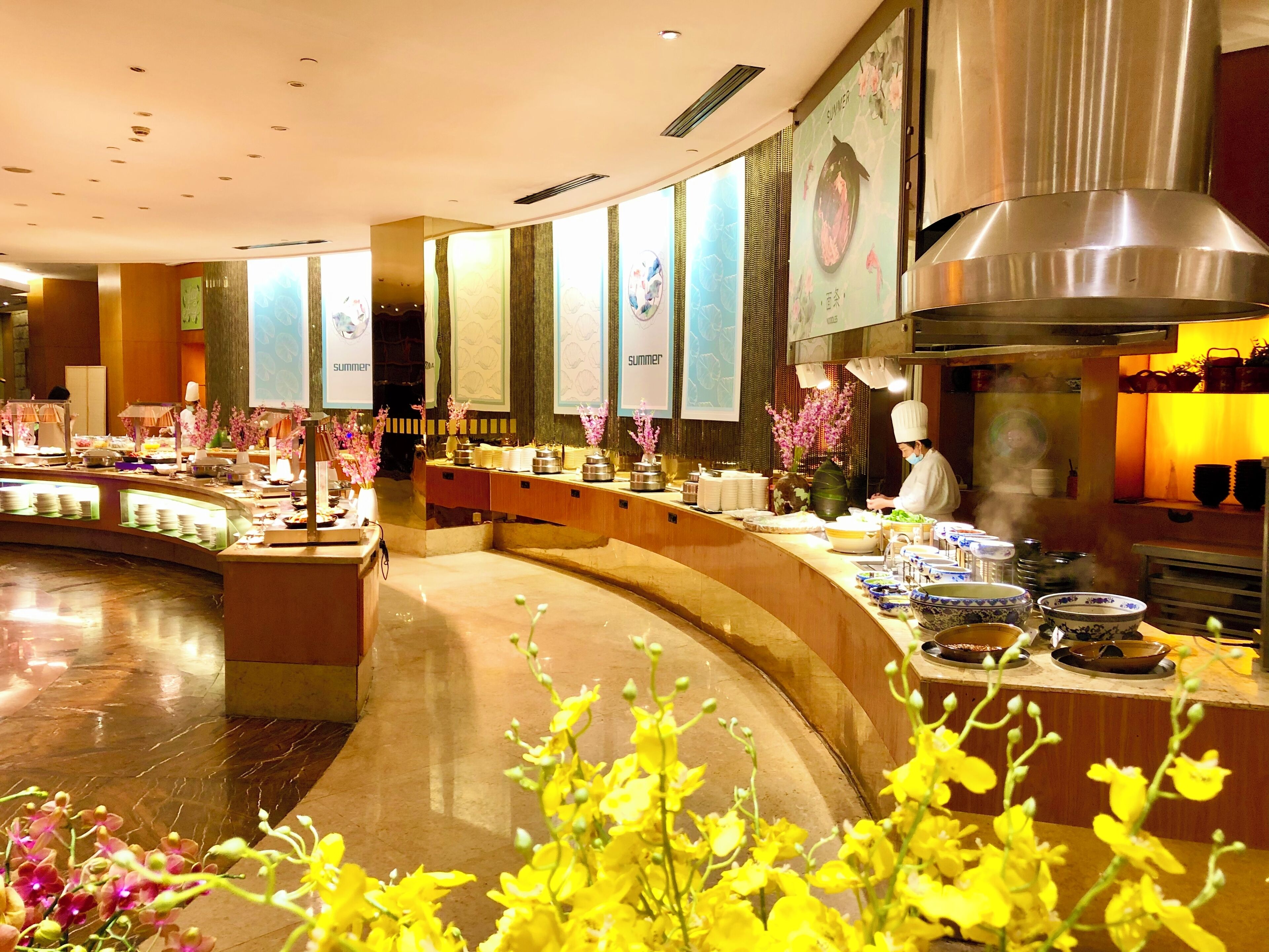 Et hotel lobby med en buet receptionsdisk, en kok der tilbereder mad, og et buffetområde med forskellige retter og blomsterdekorationer.
