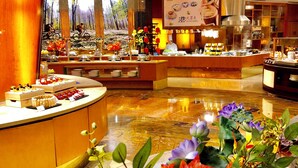 Un hall de hotel con una recepción central, un ventanal con vistas a un bosque y una zona de bufé con diversos alimentos.