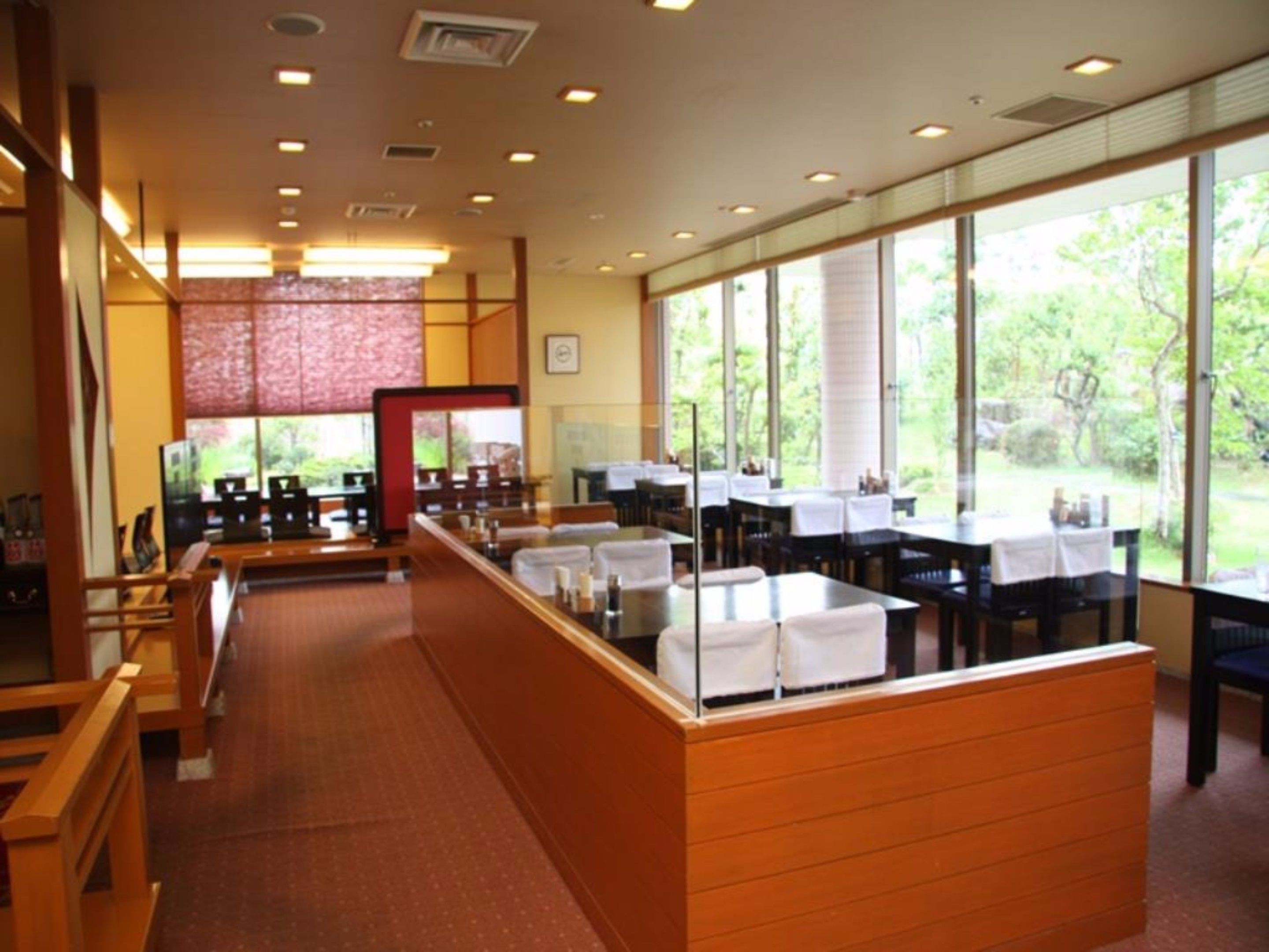 A restaurant with a long counter, multiple tables, and large windows offering a view of greenery.