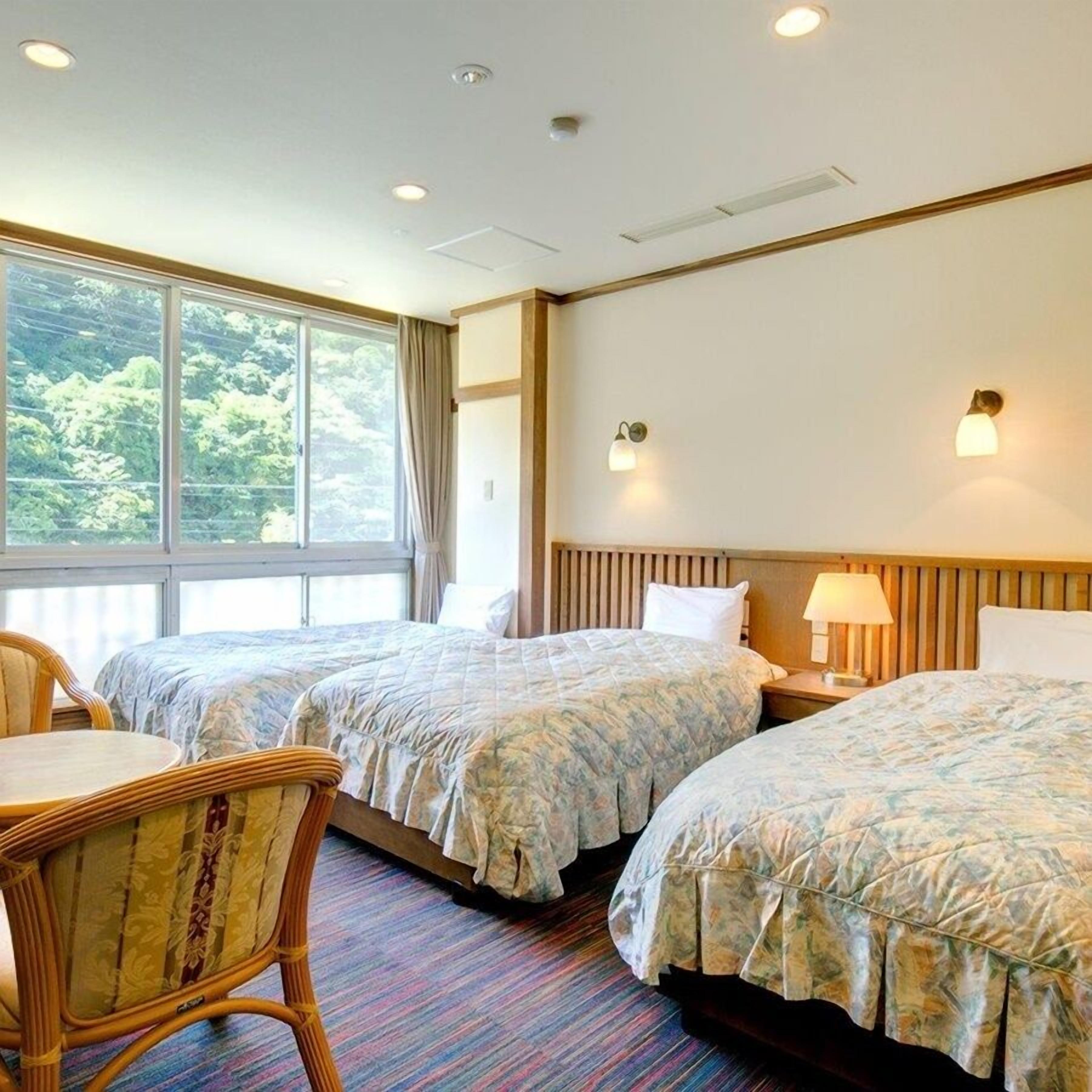 A hotel room with two beds, a window with a view of trees, a wooden headboard, and a nightstand with a lamp.