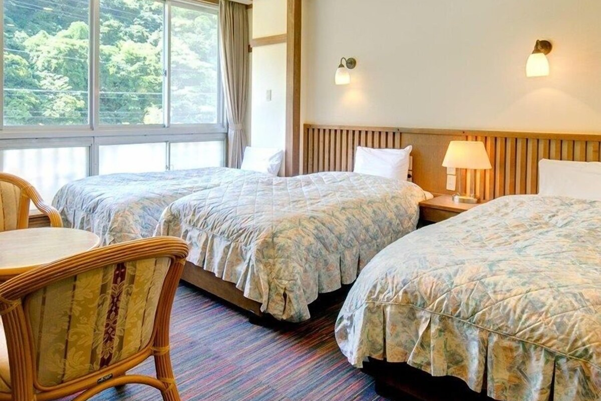 A hotel room with two beds, a window with a view of trees, a wooden headboard, and a nightstand with a lamp.