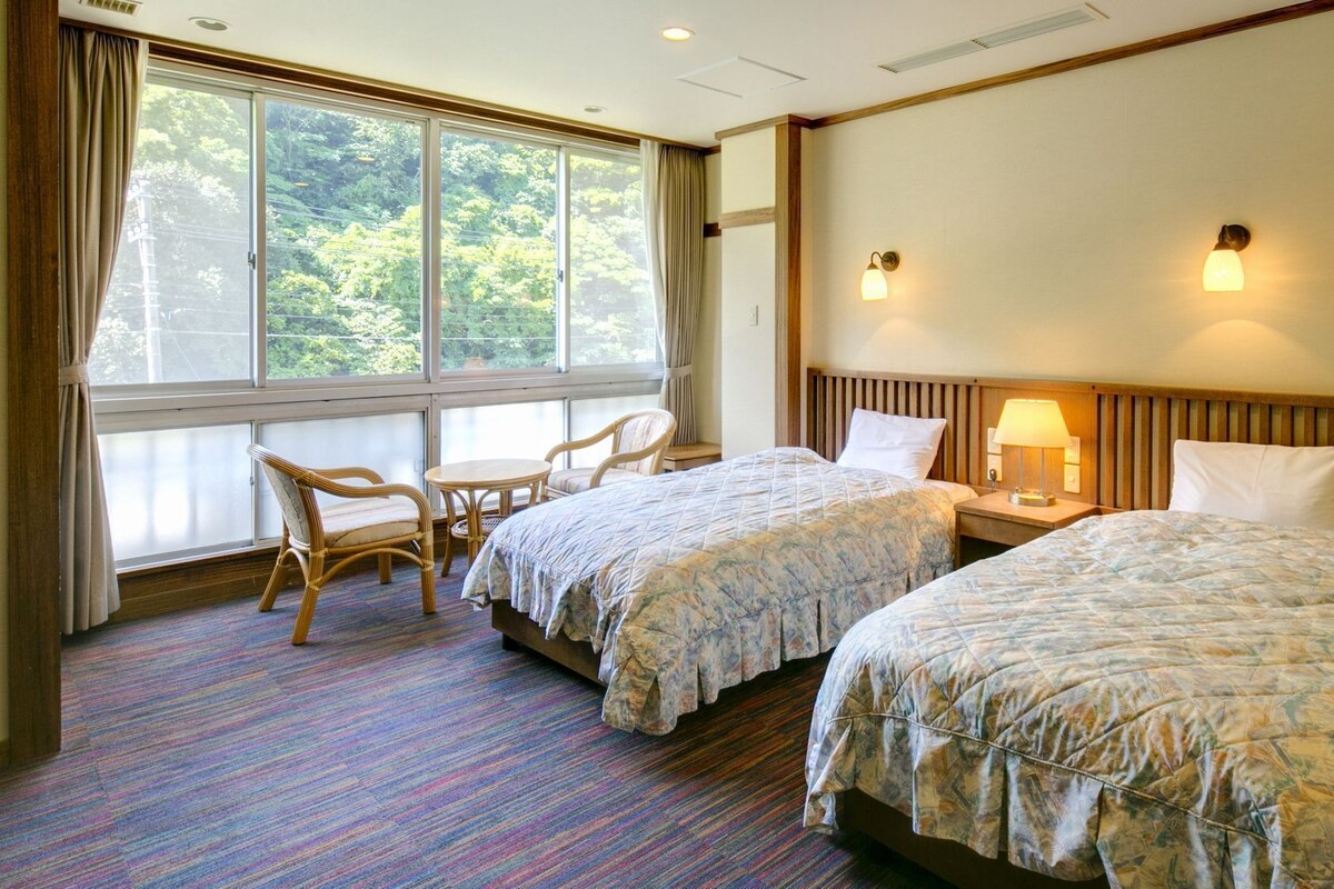 A hotel room with two beds, a small table, two chairs, a window with curtains, and a headboard with a light fixture.