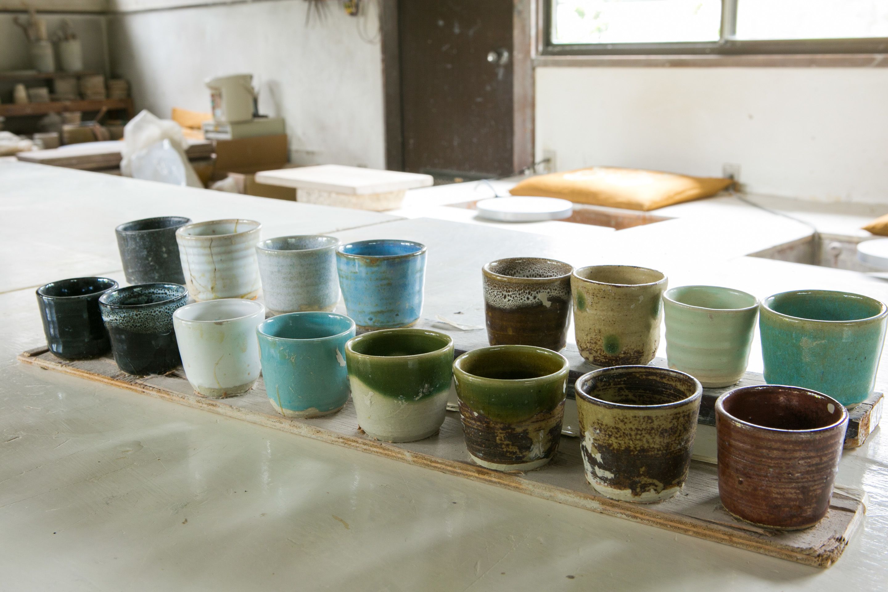 A variety of ceramic cups and bowls on a wooden tray in a workshop setting.