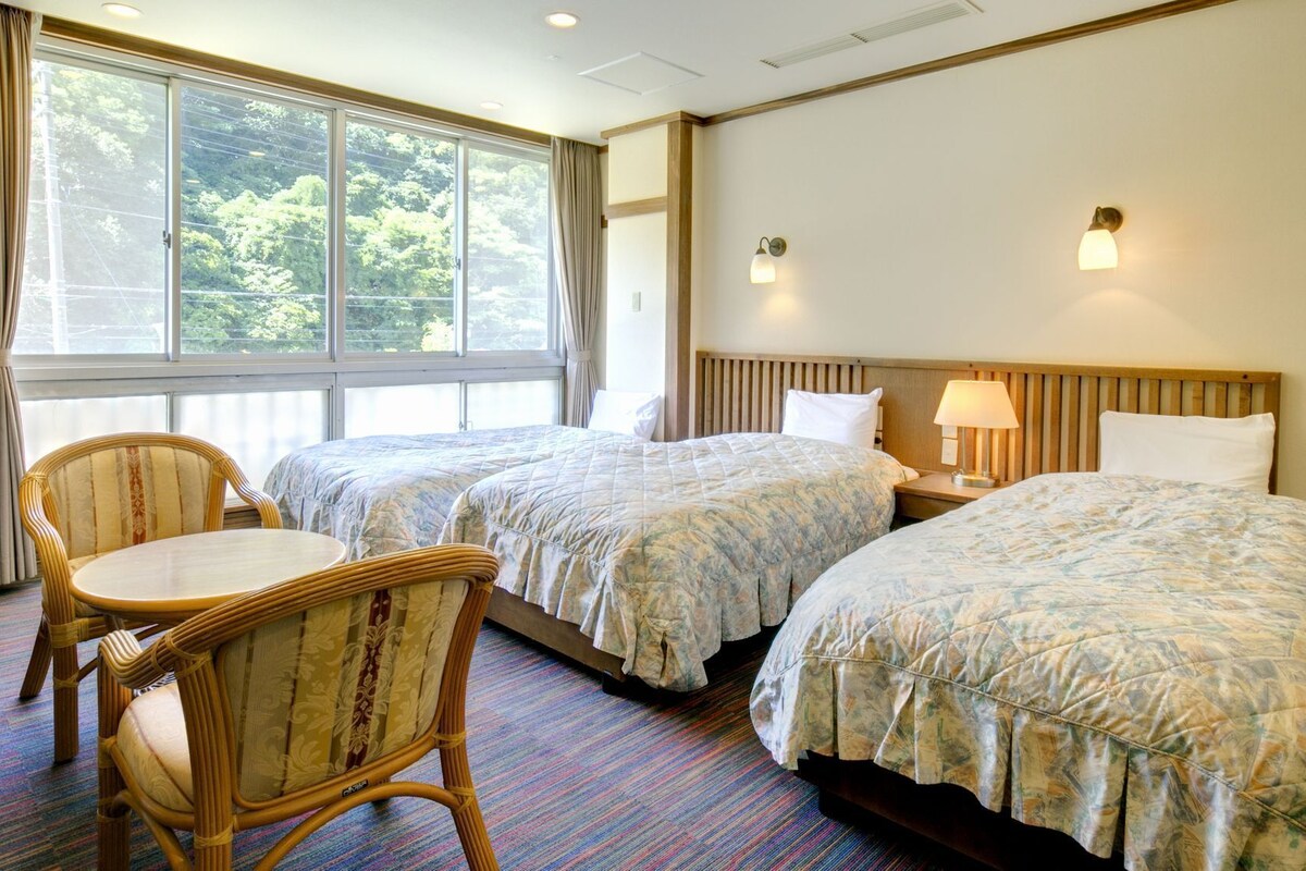 A hotel room with two beds, a chair, a window with curtains, and a lamp.