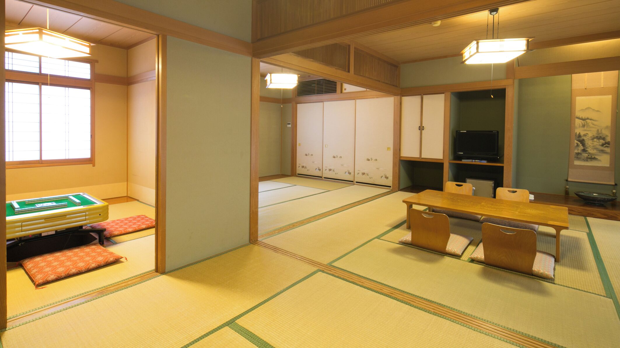 A tatami mat room with a futon bed, a low table with chairs, a television, and a painting on the wall.