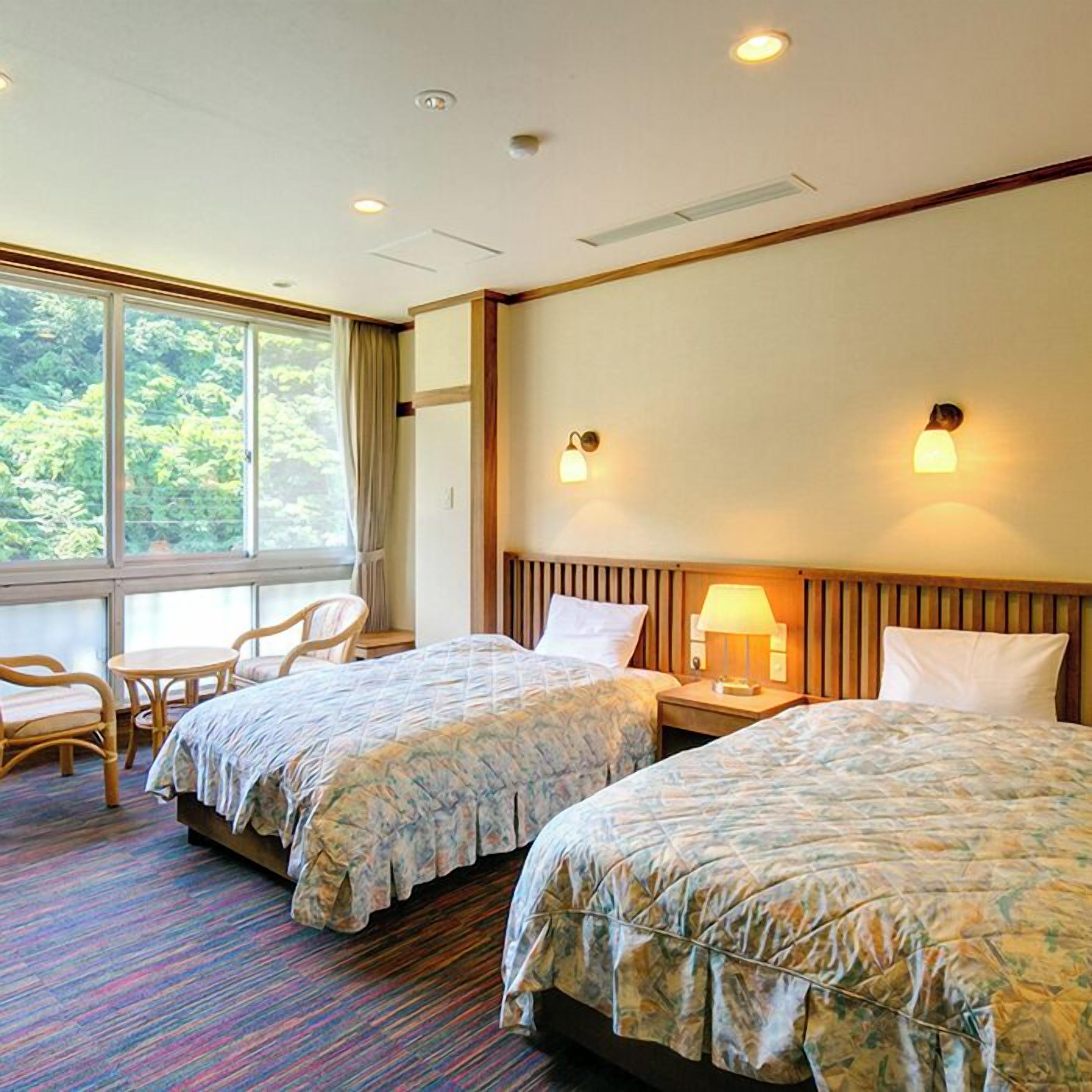 A hotel room with two beds, a small table, chairs, and a view of greenery through the window.