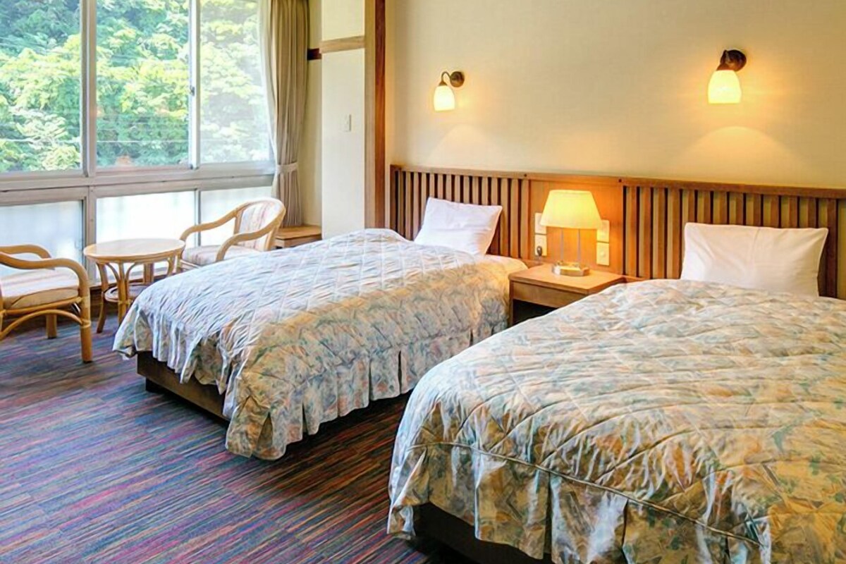 A hotel room with two beds, a small table, chairs, and a view of greenery through the window.