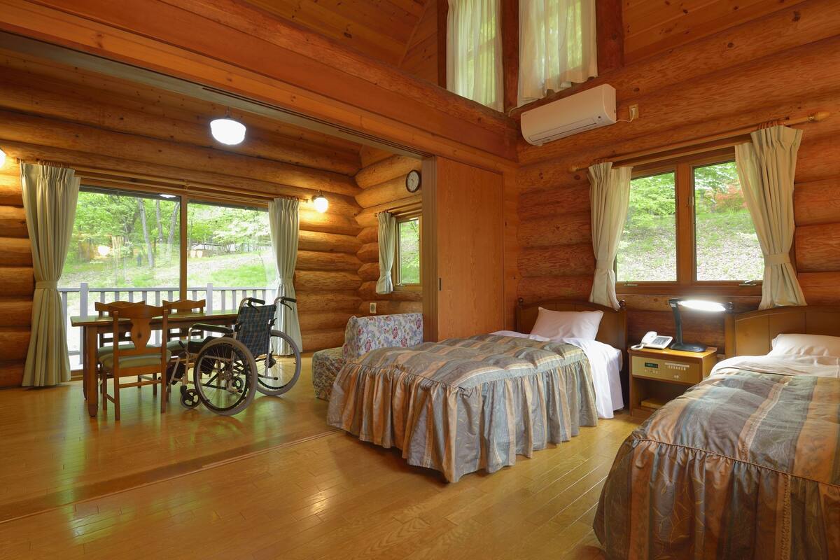 A log cabin room with two beds, a wheelchair, a table with chairs, and a view of the outdoors.