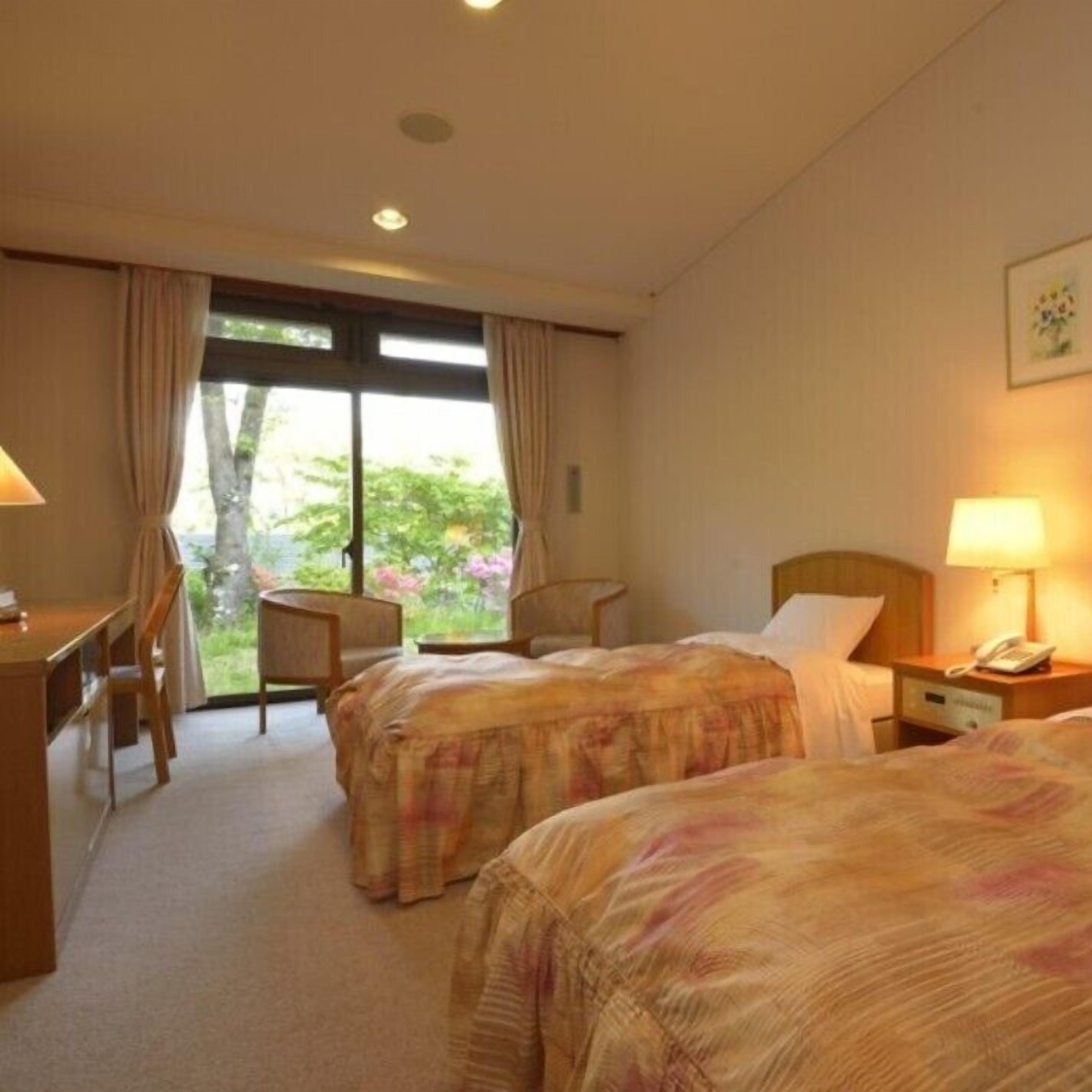 A hotel room with two beds, a desk, a chair, and a window with a view of greenery.