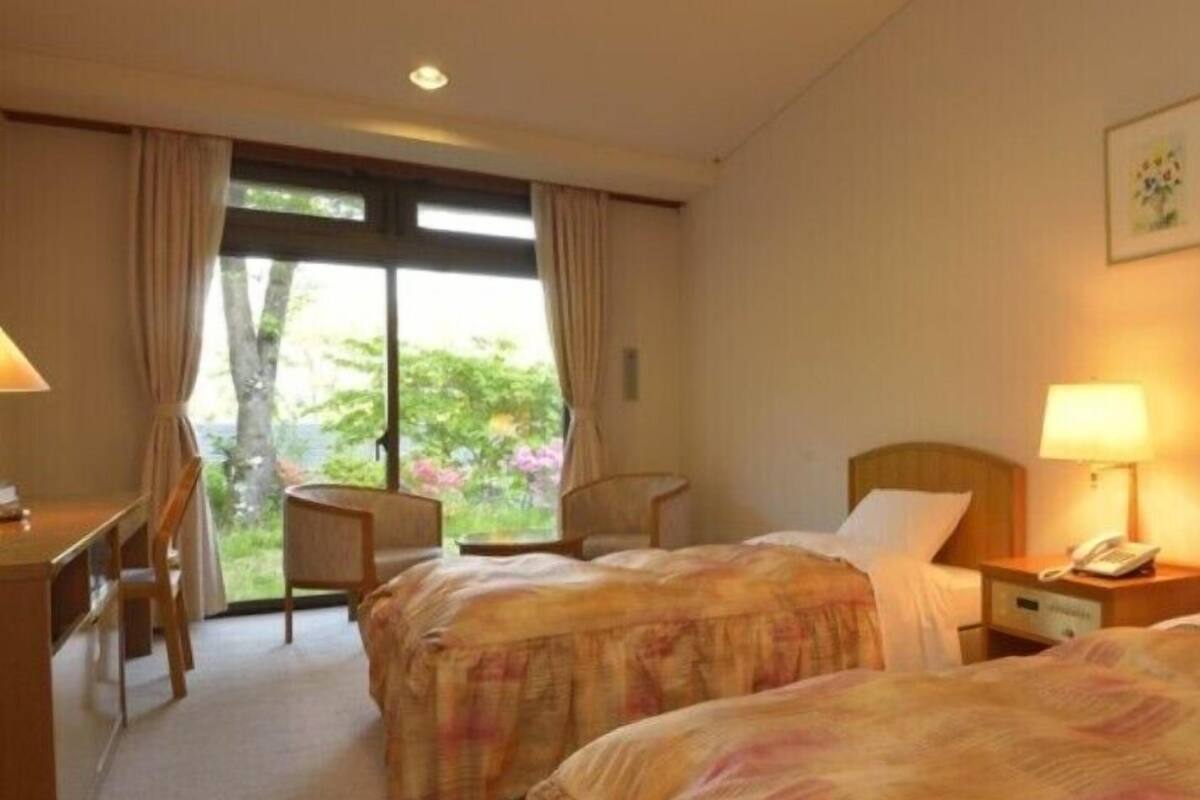 A hotel room with two beds, a desk, a chair, and a window with a view of greenery.