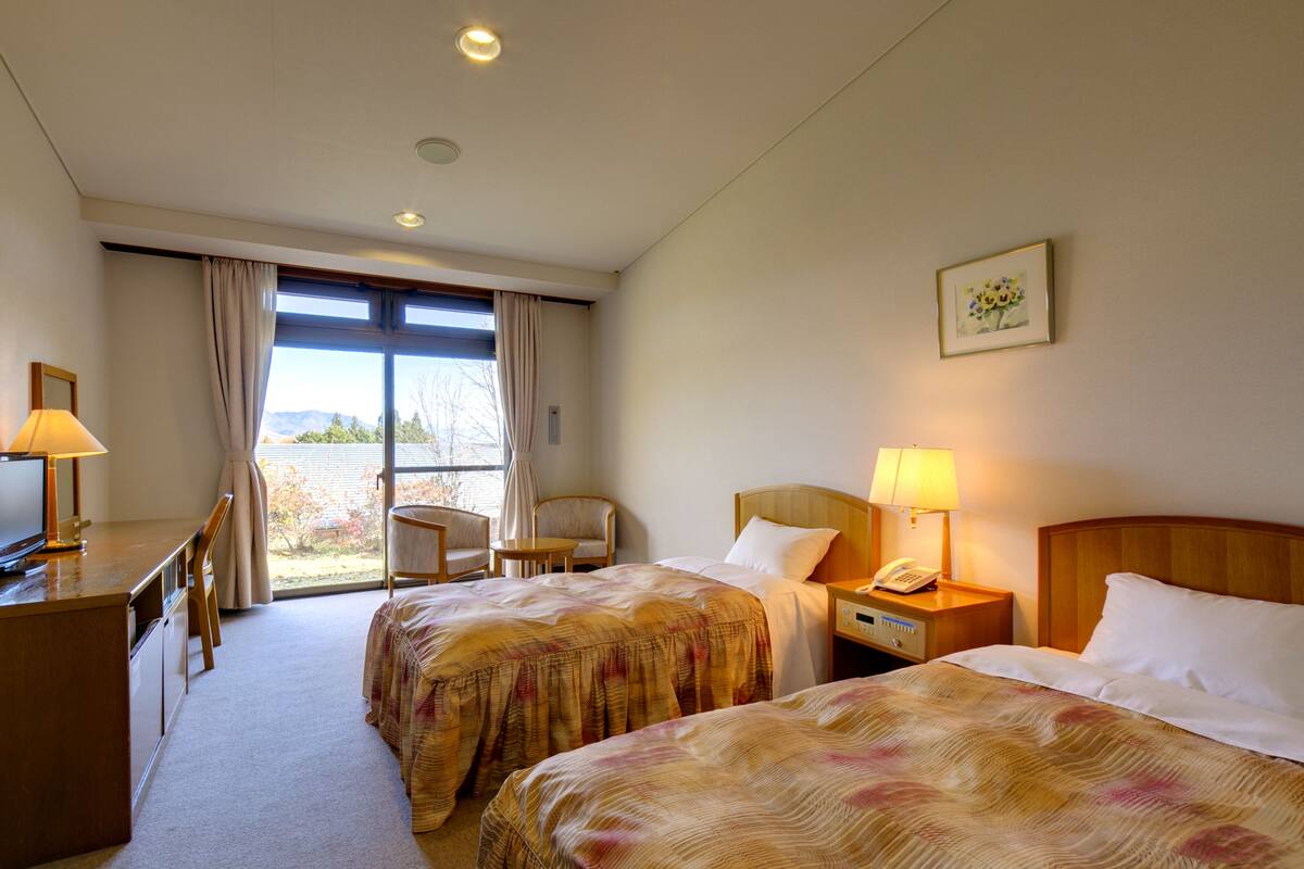 A hotel room with two beds, a desk, a chair, a small table, and a window with curtains.