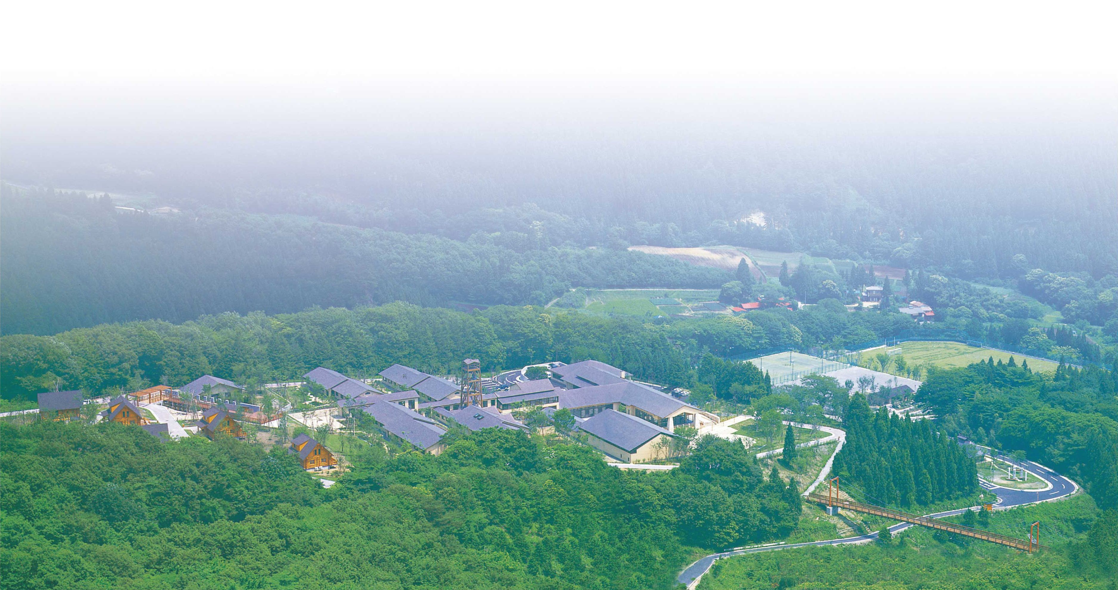 An aerial view of a resort with multiple buildings surrounded by dense forest, a winding road, and a distant view of a hilly landscape.