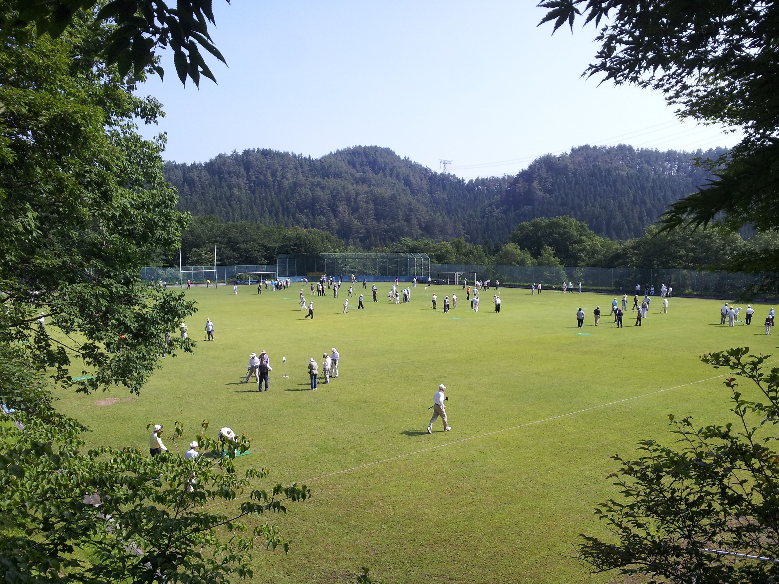 Um extenso campo gramado e aberto com pessoas espalhadas, rodeado por árvores e colinas ao fundo.