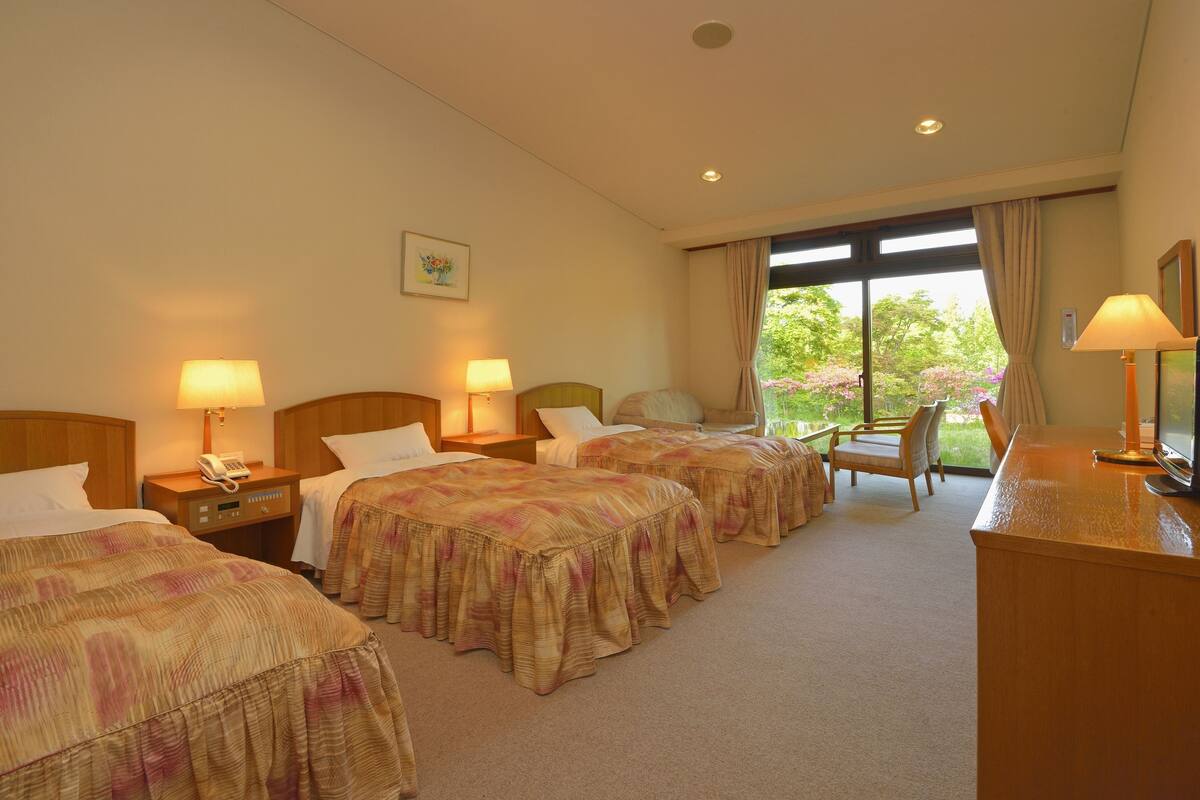 A hotel room with two beds, a desk, a chair, and a window with a view of greenery.
