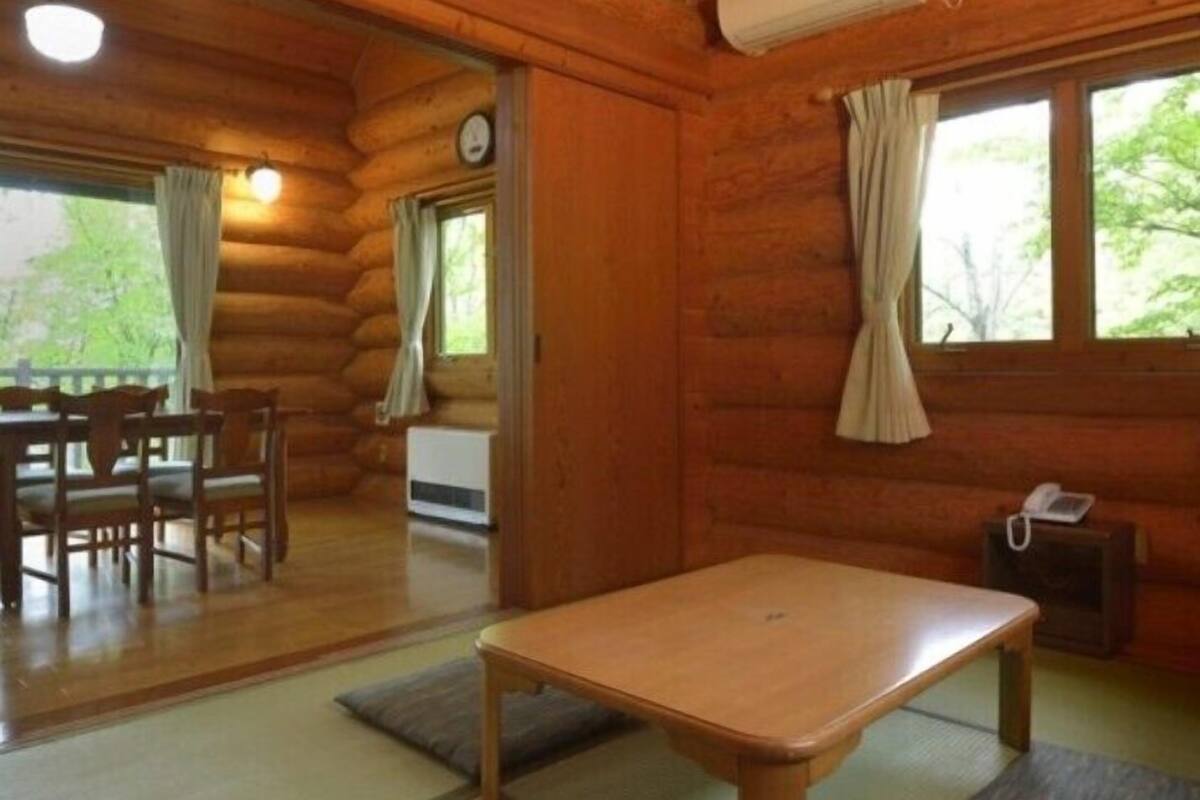 A log cabin interior with a dining table, chairs, a small sofa, and a window with curtains.