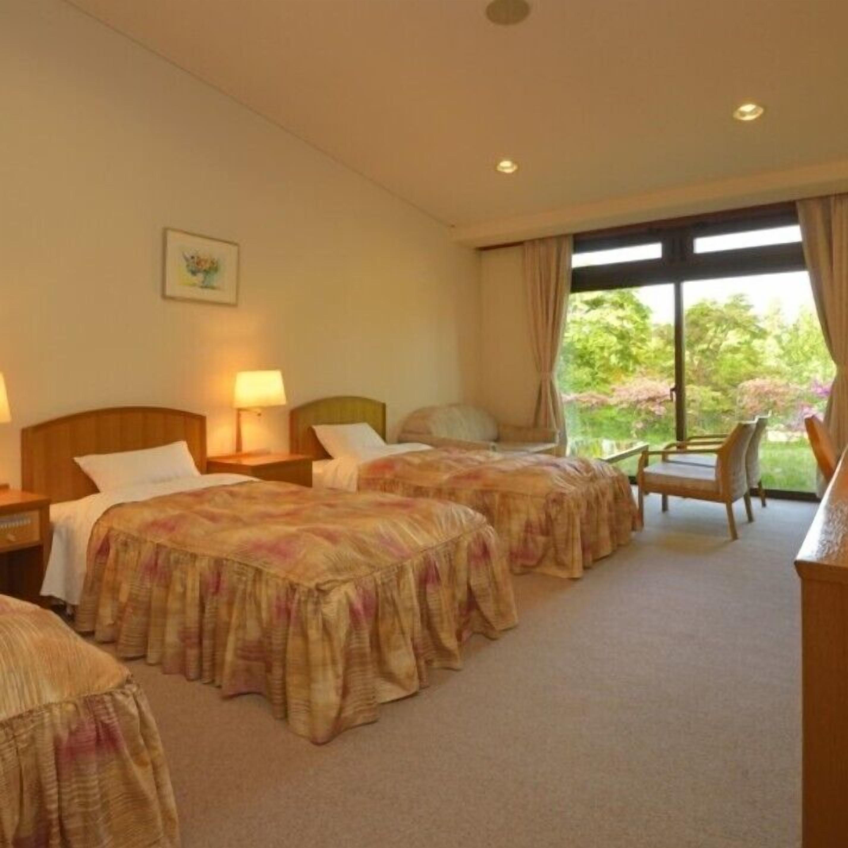 A hotel room with two beds, a desk, a chair, and a window with a view of greenery.