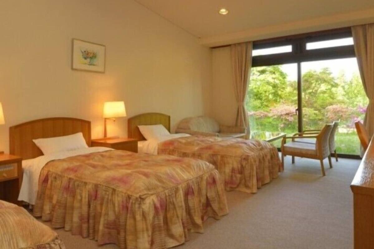 A hotel room with two beds, a desk, a chair, and a window with a view of greenery.