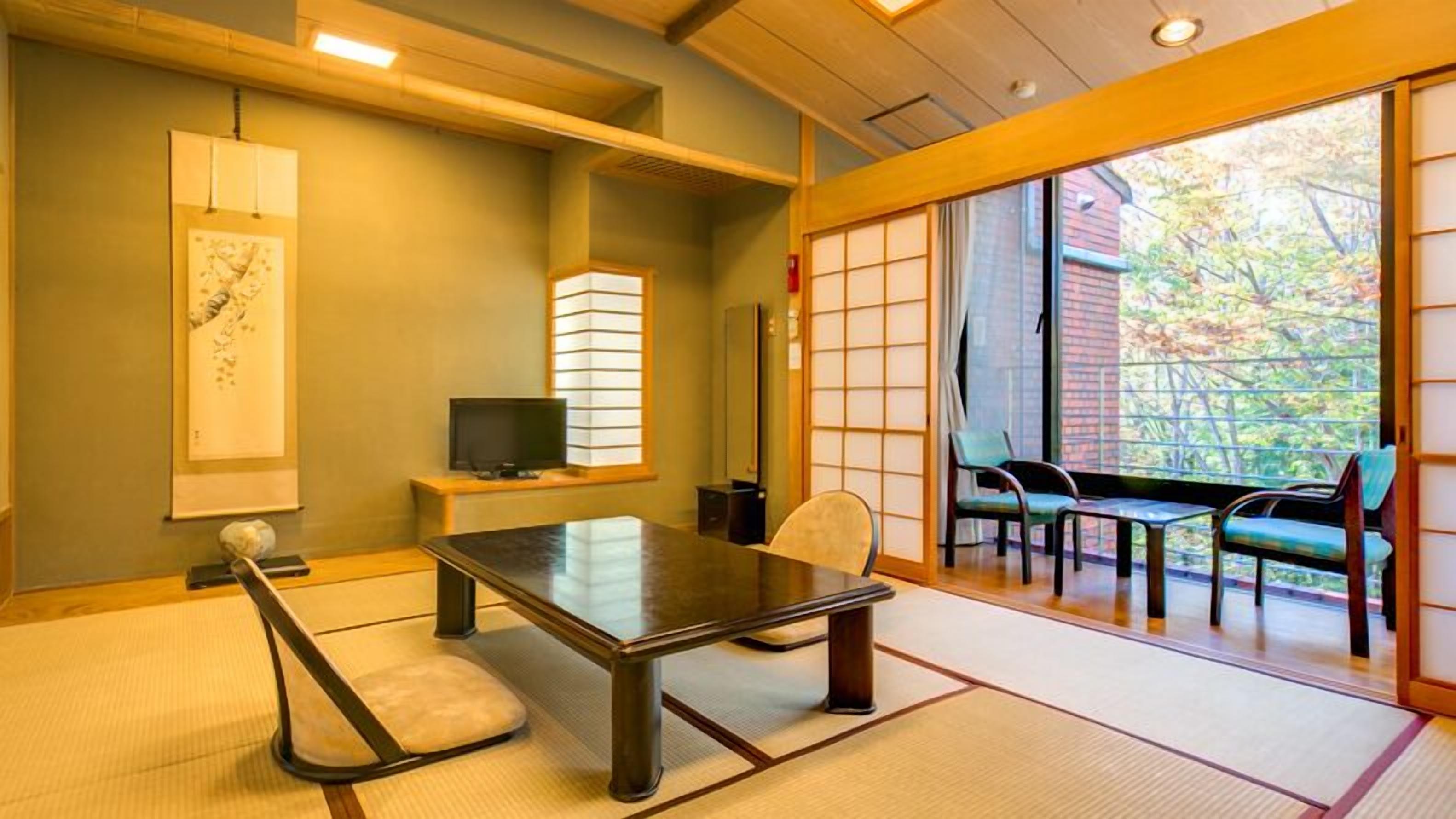 A traditional Japanese room with tatami flooring, a low table, a television, and sliding doors leading to a balcony with a view of trees.