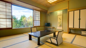 Una habitación con suelo de tatami, una mesa baja y sillas, un televisor en un estante y puertas correderas.