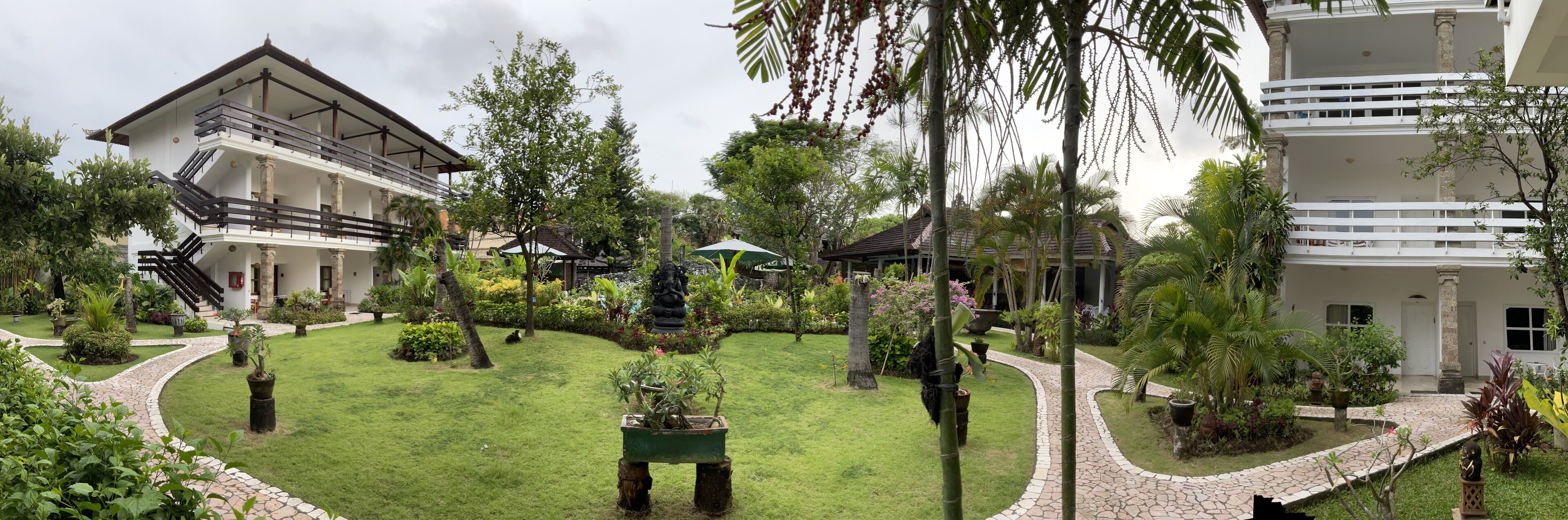 Un patio con una estatua central, rodeado de exuberante vegetación y un sendero que lleva a un edificio con varios balcones.