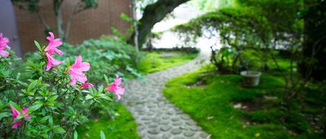A garden pathway leading to a brick building, surrounded by lush greenery and pink flowers.