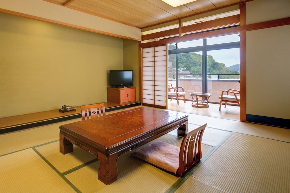 Een traditionele Japanse kamer met tatami vloeren, een lage houten tafel, een televisie op een kast en schuifdeuren die uitkomen op een balkon.