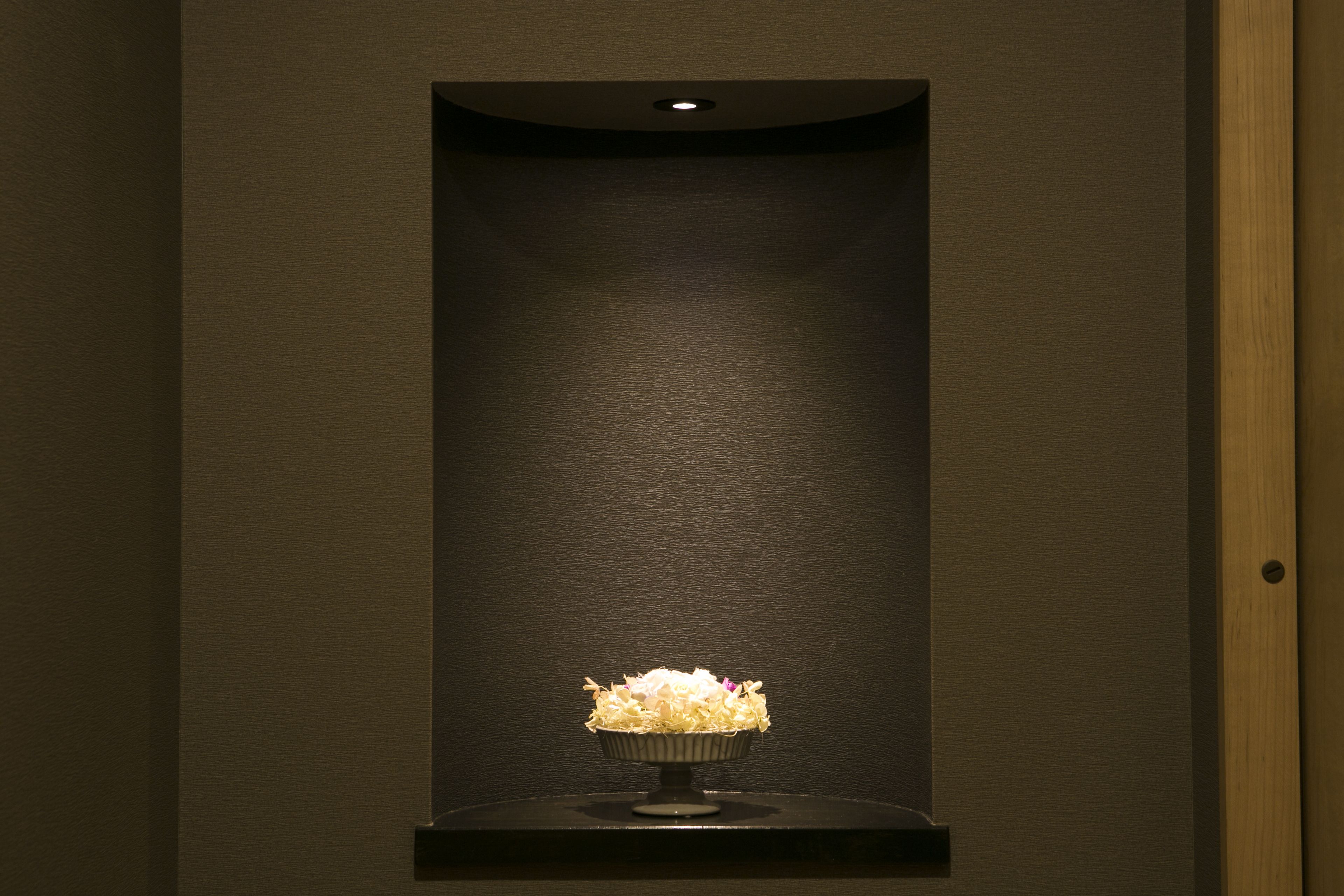 A dark room with a single light fixture on the ceiling and a small, dark shelf with a bowl of flowers.