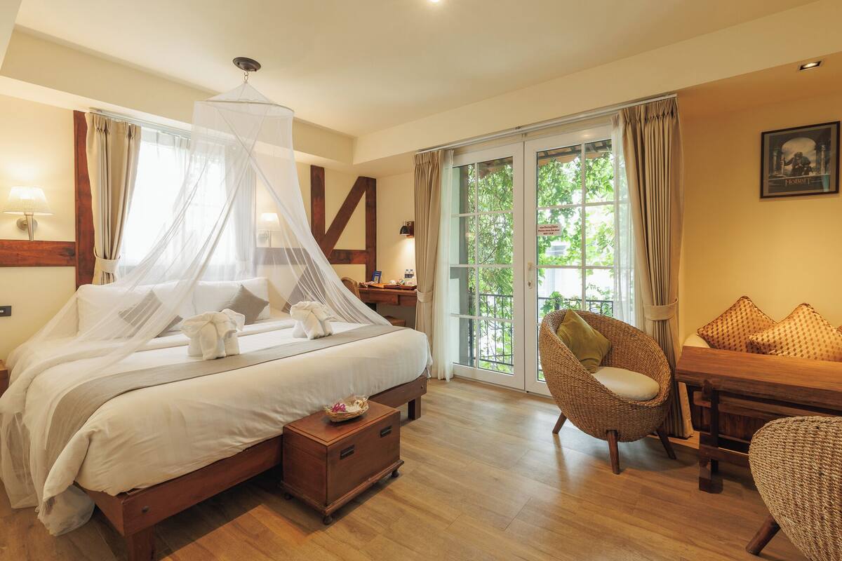 A bedroom with a four-poster bed, a sitting area with a chair and table, and a view of greenery through the window.