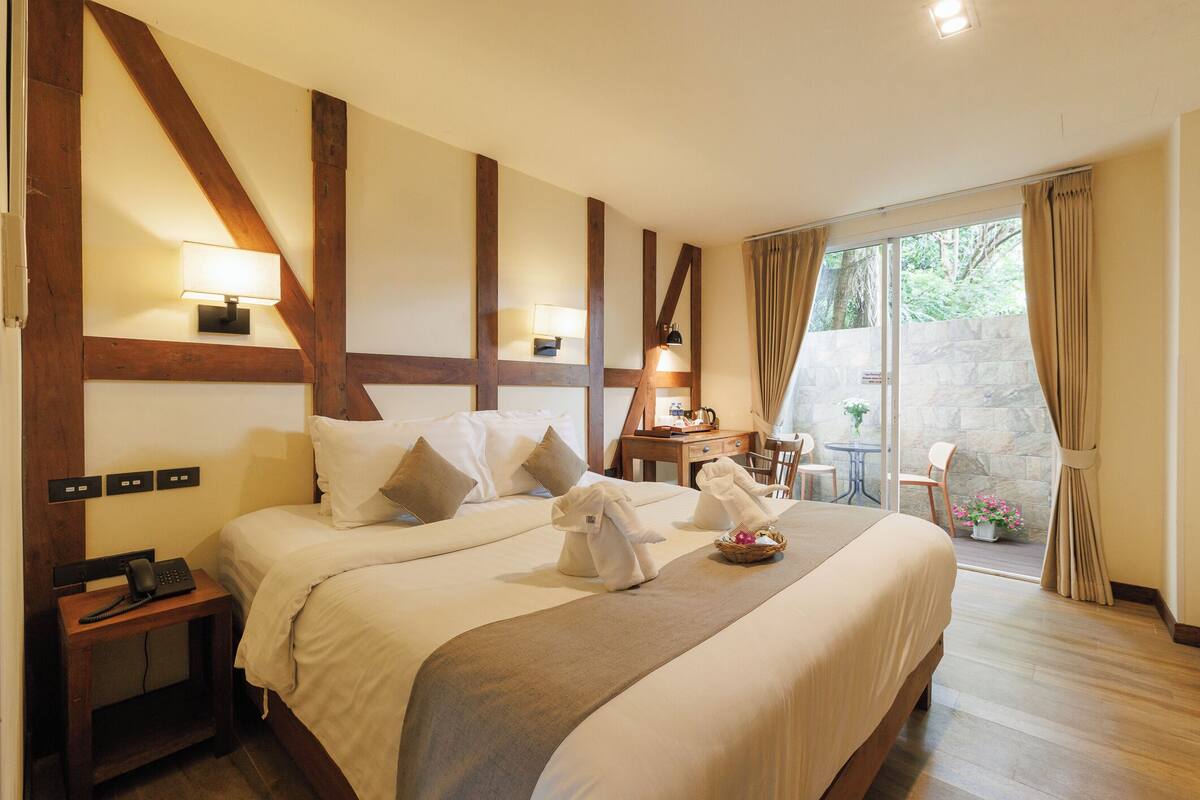 A hotel room with a large bed, bedside lamps, a telephone, a small table with a lamp, and a view of the outdoors through a sliding glass door.