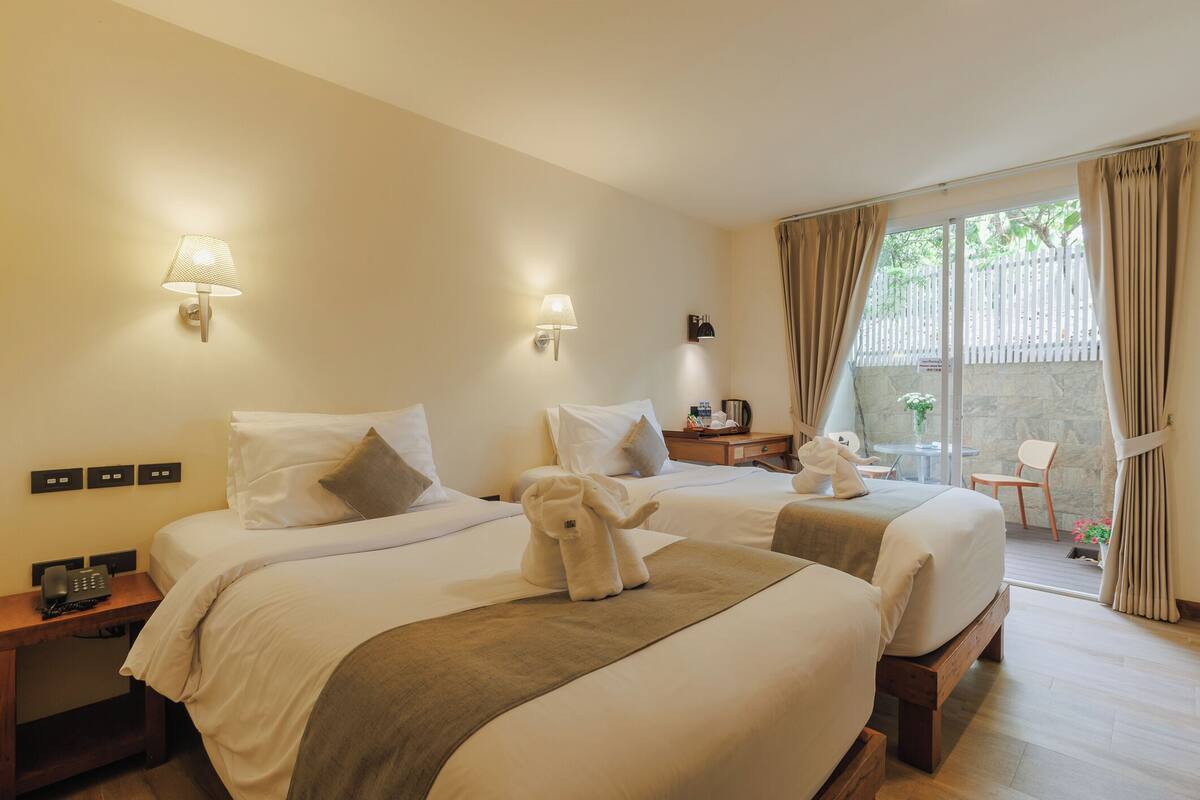 A hotel room with two beds, a small table, a chair, and a balcony with curtains.
