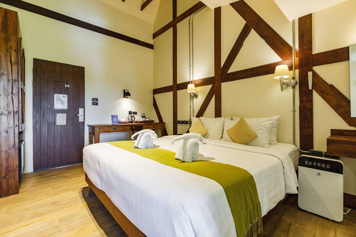 A hotel room with a large bed, wooden beams on the ceiling, a desk with a lamp, and a suitcase.