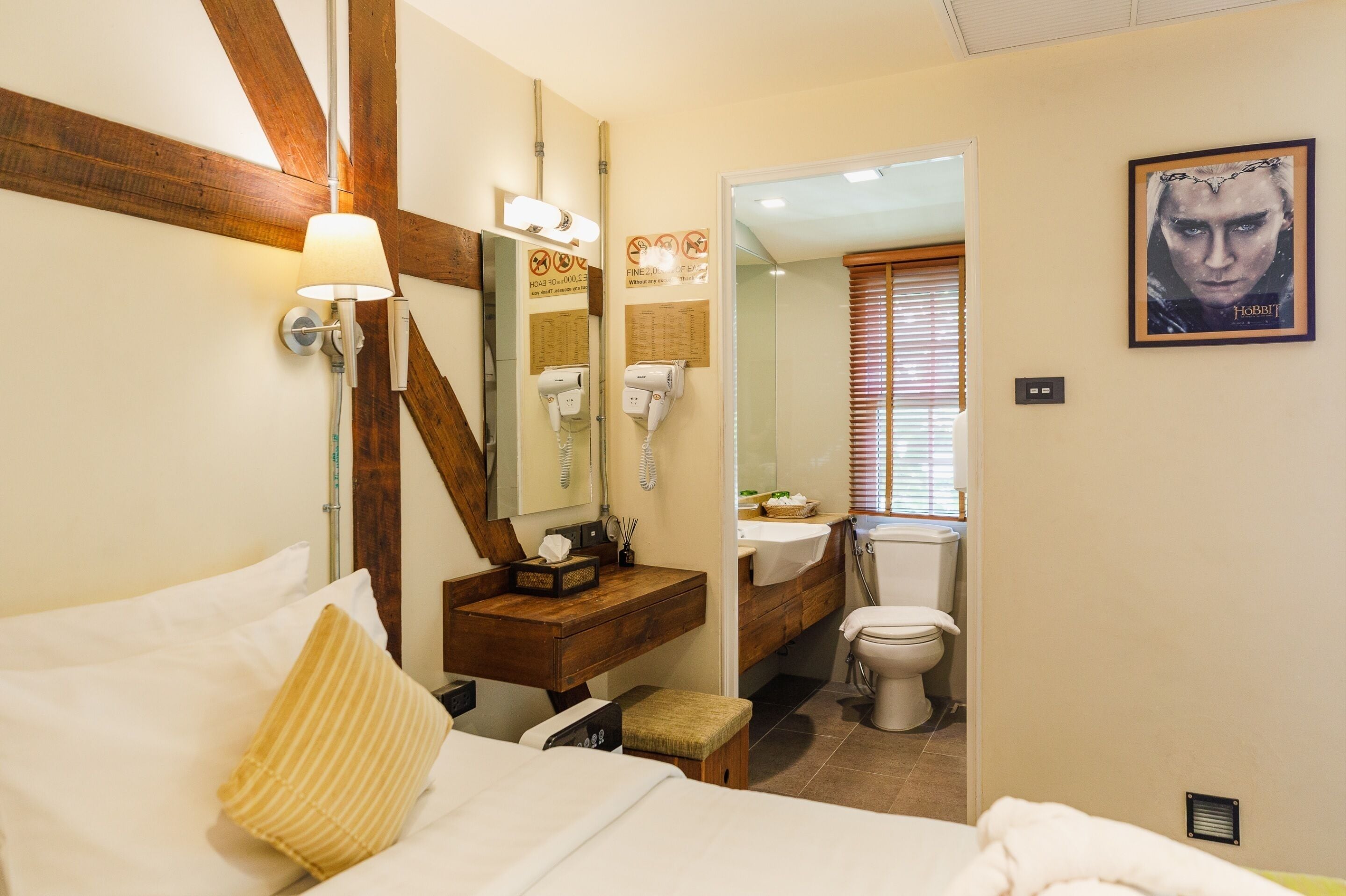 A hotel room with a bed, a wooden headboard, a bedside table with a lamp, a mirror, a bathroom with a toilet and sink, and a framed picture on the wall.