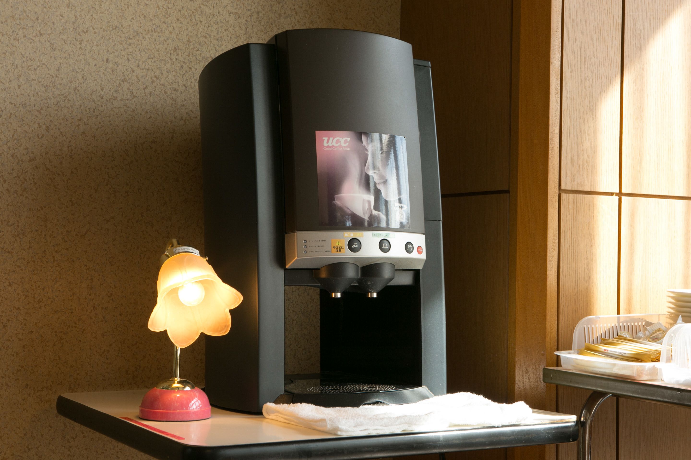 A coffee machine with a digital display, a table with a lamp, and a stack of plates.