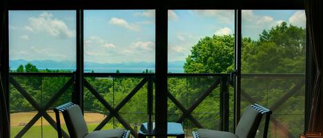 A room with large windows offering a view of a baseball field and mountains. There are two chairs and a small table in the room.