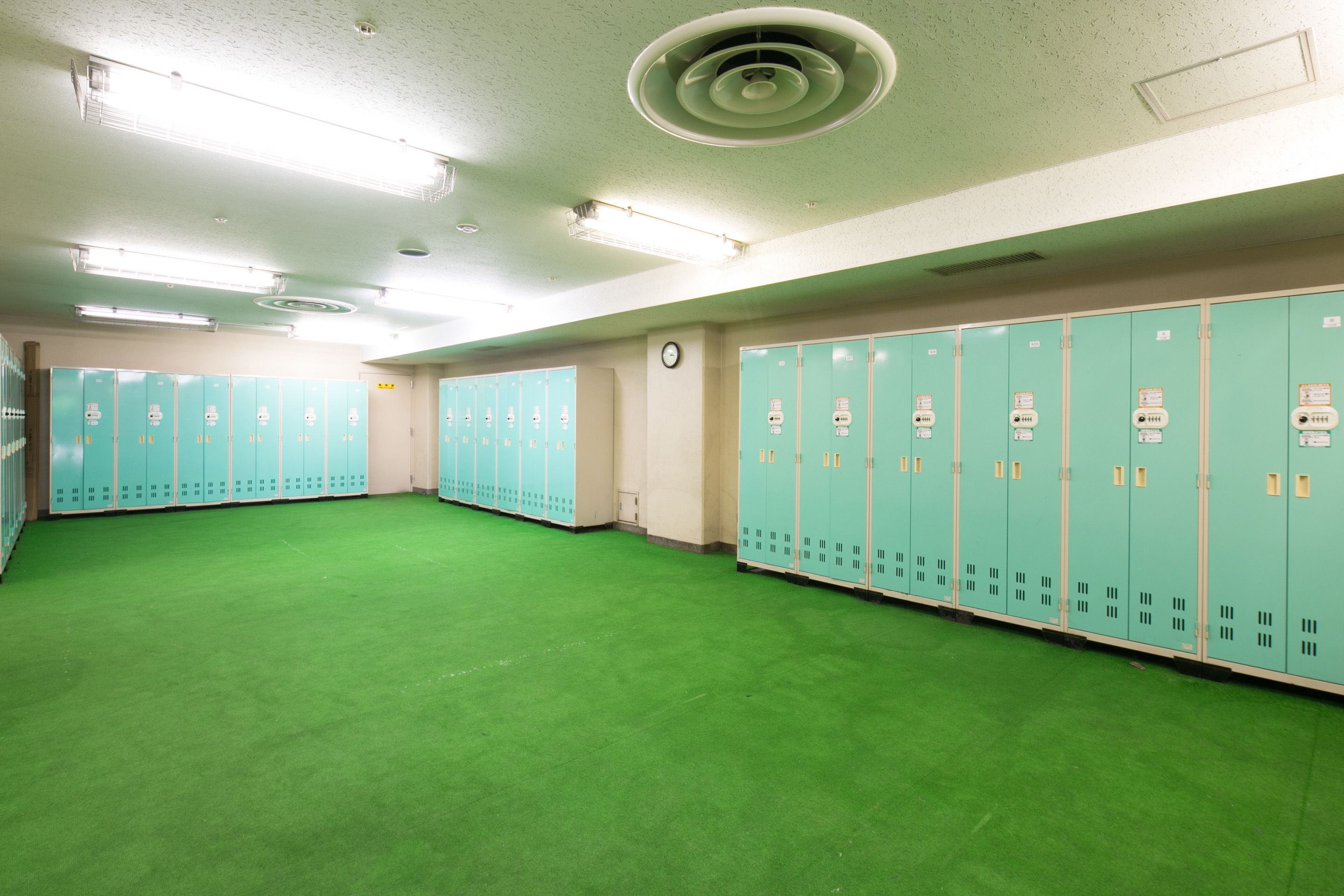 A locker room with green lockers, a clock on the wall, and a green carpeted floor.