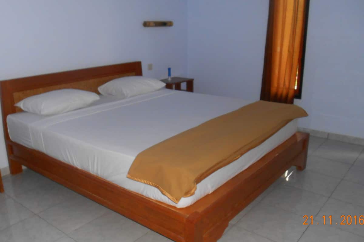 A standard hotel room with a wooden bed, white linens, and a brown throw blanket. There is a nightstand with a lamp and a small table beside the bed.