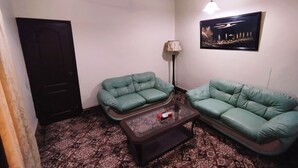 A living room with two green sofas, a patterned carpet, a ceiling fan, a lamp, and a framed picture on the wall. - Hilltop Hotel (Karachi)
