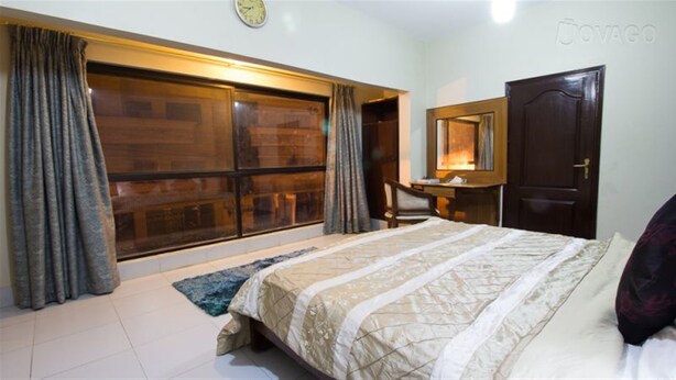 A bedroom with a large bed, a desk, a chair, a wardrobe, and a window with curtains. - Hilltop Hotel (Karachi)