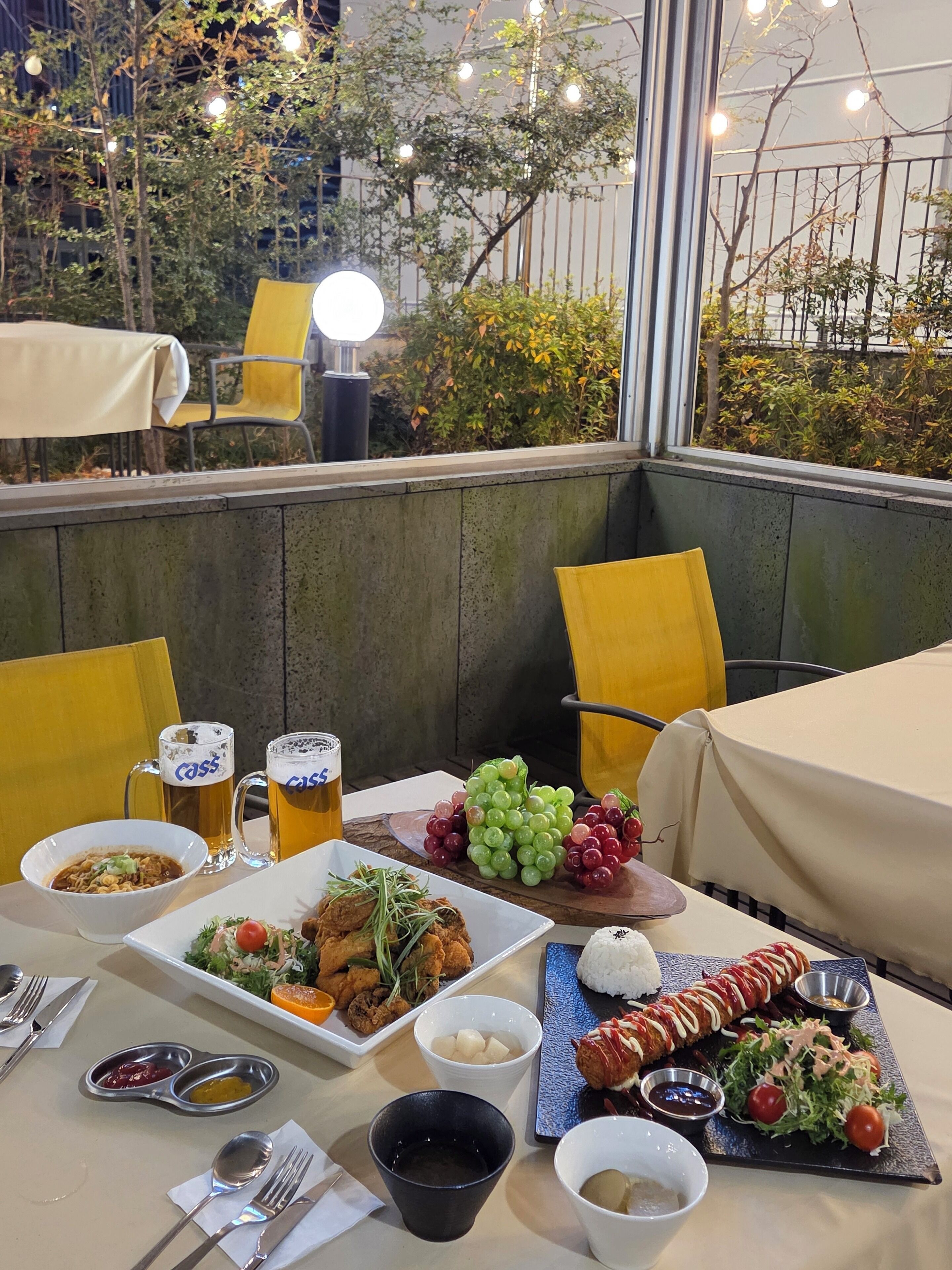 An outdoor dining area with a table set for two, featuring a variety of dishes, drinks, and a scenic view of trees and lights.