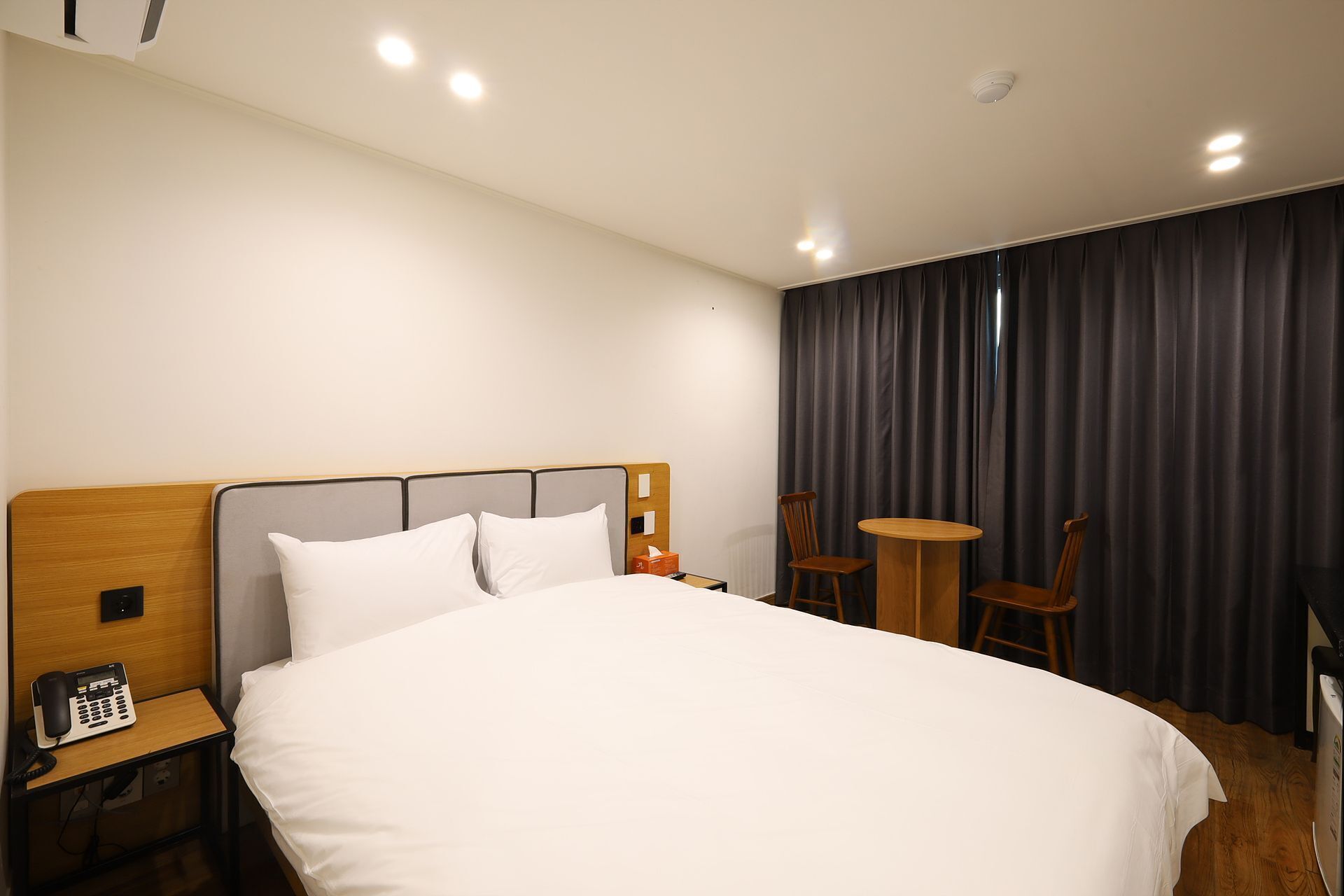 A hotel room with a large bed, a bedside telephone, a small table with two chairs, and a window with dark curtains.