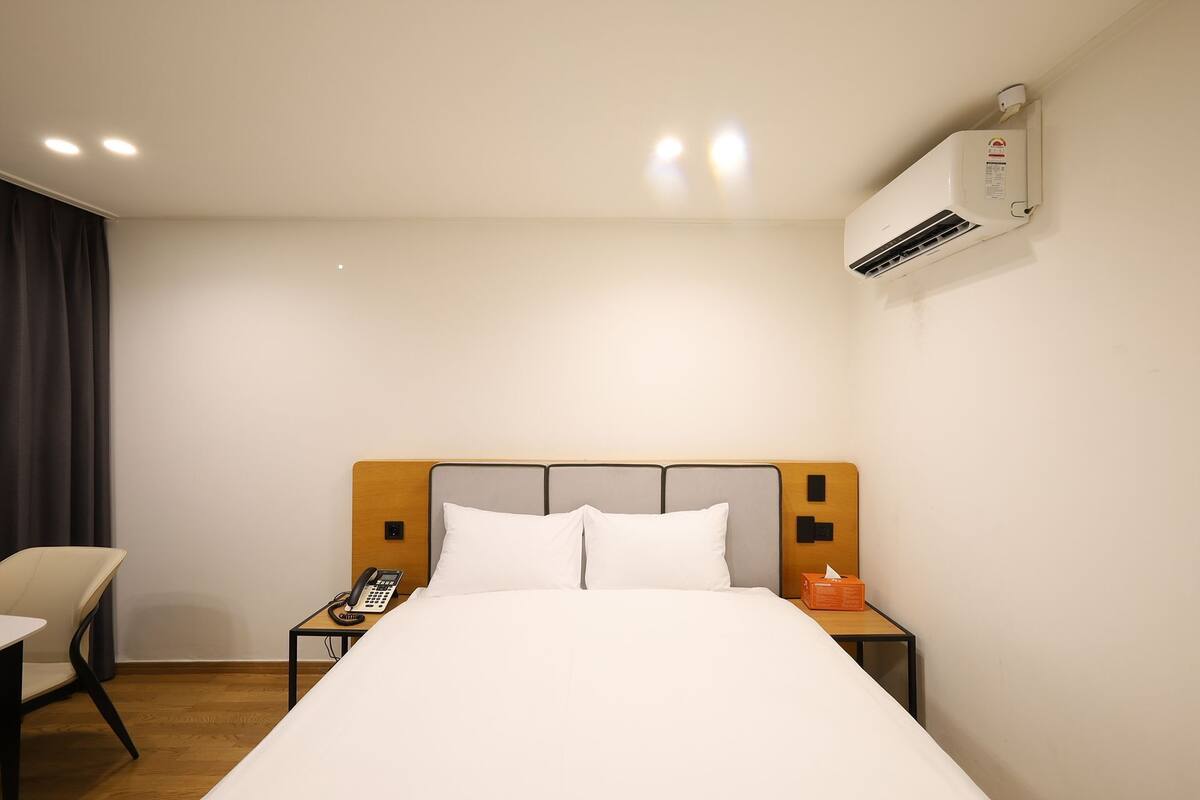 Habitación de hotel con cama, mesita de noche con teléfono, una silla y un equipo de aire acondicionado en la pared.