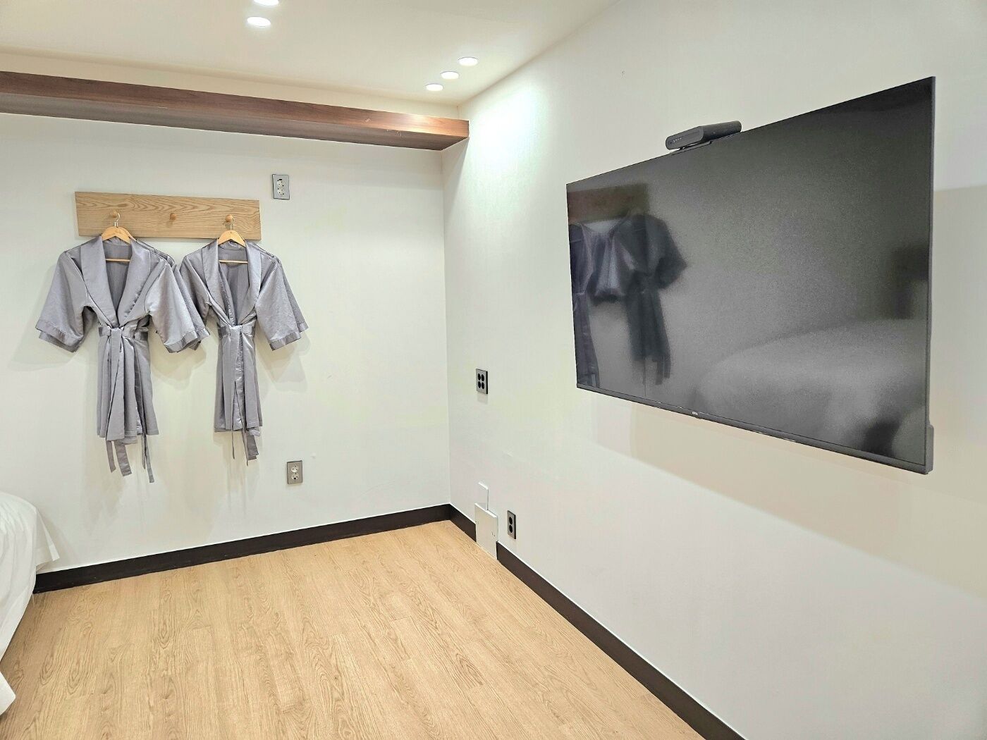 A hotel room with a large flat-screen TV mounted on the wall, wooden flooring, and two robes hanging on a wooden rack.