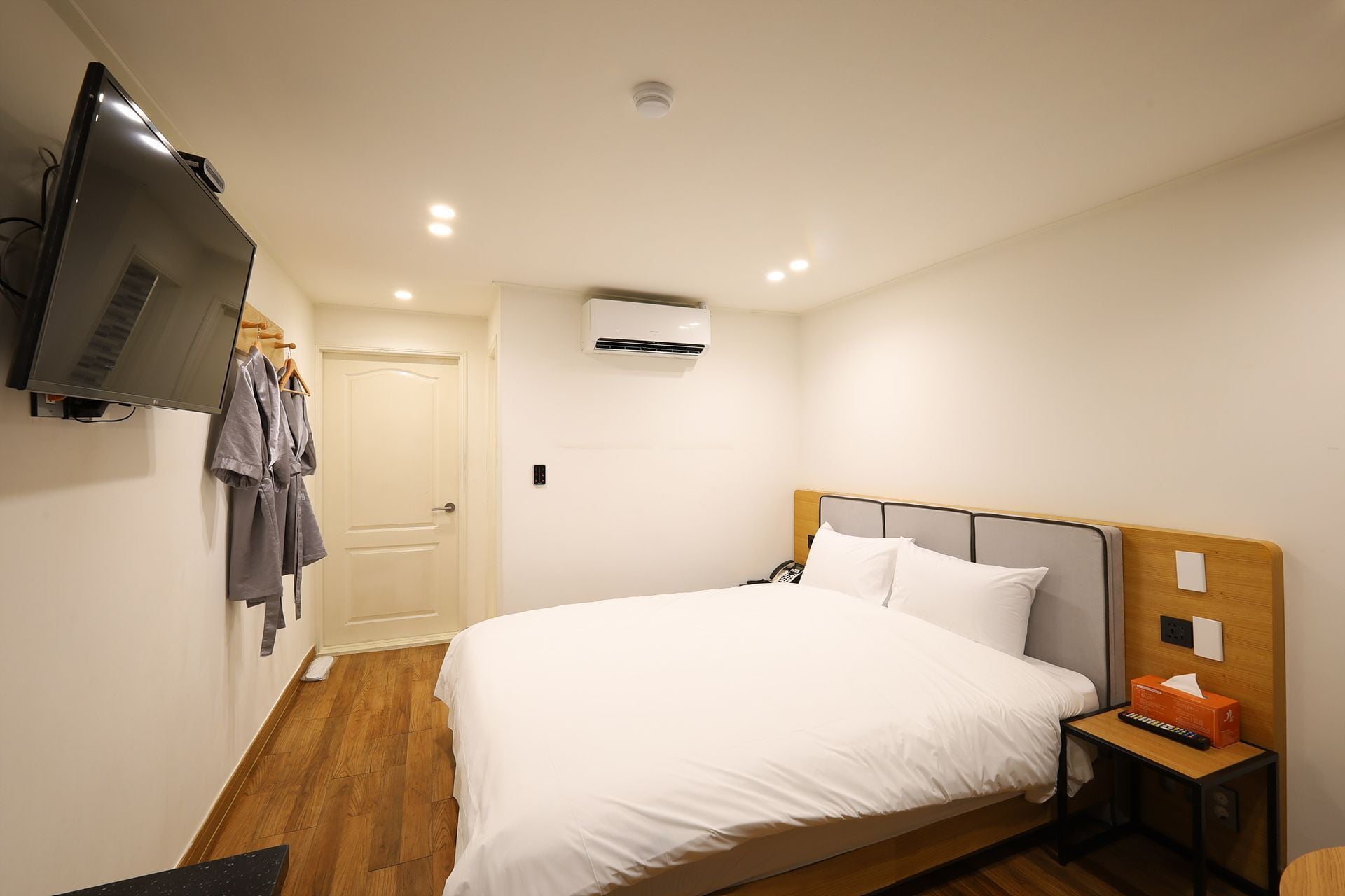 A hotel room with a bed, a TV mounted on the wall, an air conditioning unit, and a wooden floor.