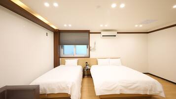 A hotel room with two beds, a window with blinds, an air conditioning unit, and a bedside table with a lamp.