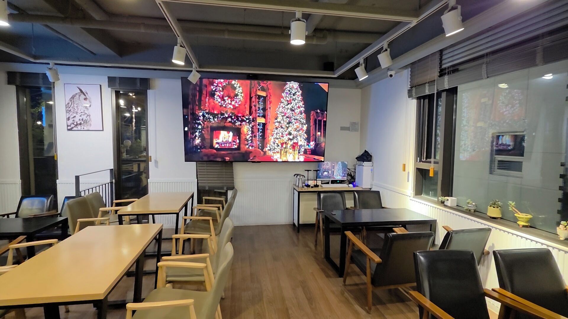 Un salón de conferencias moderno con mesas y sillas de madera, un televisor de pantalla plana grande que muestra una escena festiva, y una ventana con persianas.