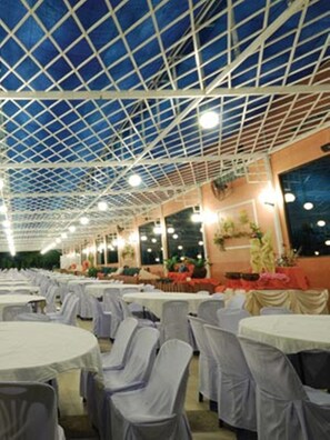 A large indoor venue with a high ceiling and a grid-like structure. Tables are arranged in rows with chairs covered in white. - R.S. Hotel (Kanchanaburi)
