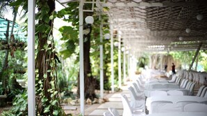 A covered outdoor seating area with white chairs and tables, surrounded by greenery and a pergola structure. - R.S. Hotel (Kanchanaburi)