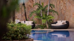 A pool area with two wicker lounge chairs, one with white cushions and one with beige cushions, surrounded by lush greenery and a stone wall. - R.S. Hotel (Kanchanaburi)