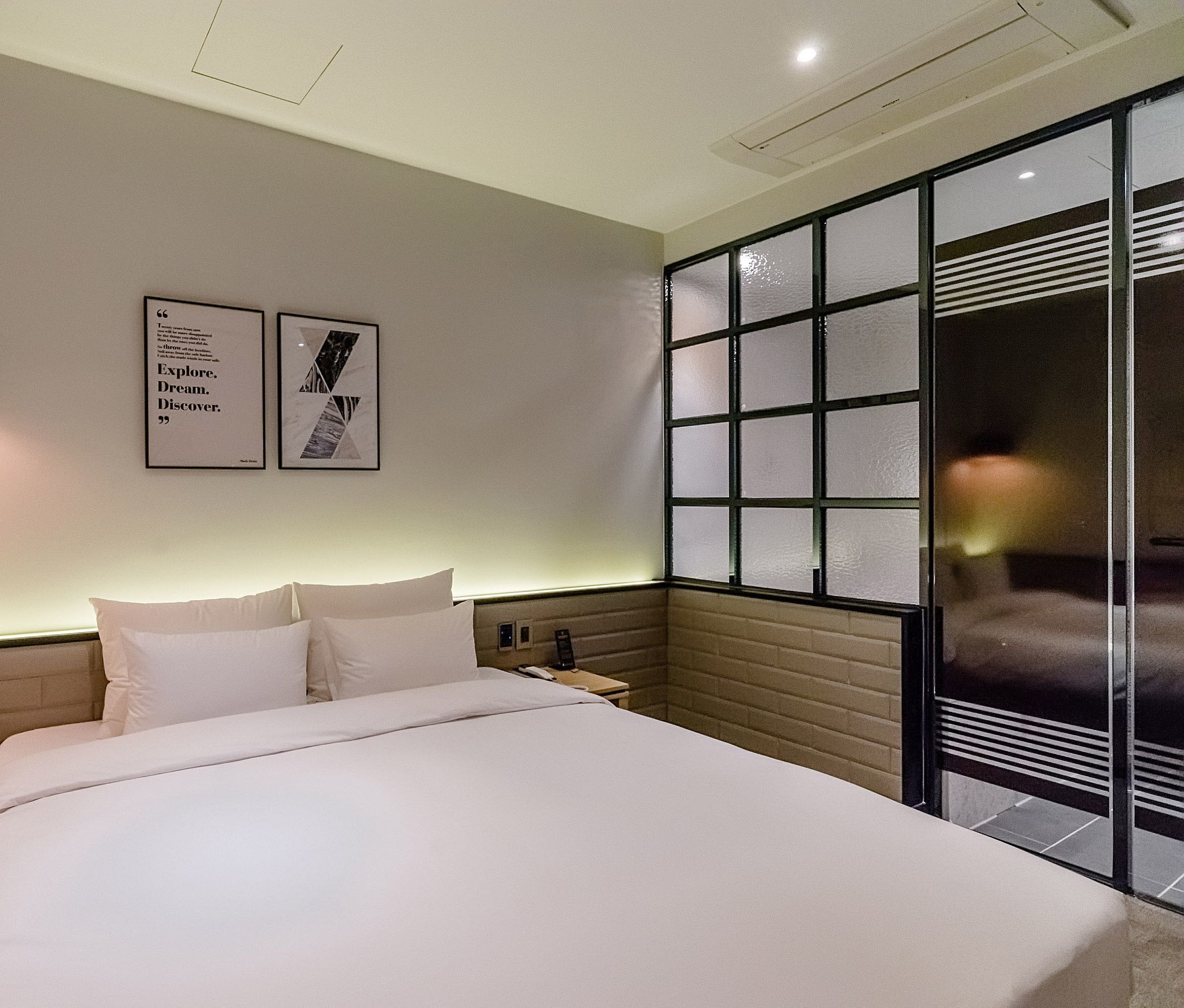 A modern hotel room with a large bed, a nightstand, and a wall-mounted light. There are two framed quotes on the wall and a large window with a view of the outside.
